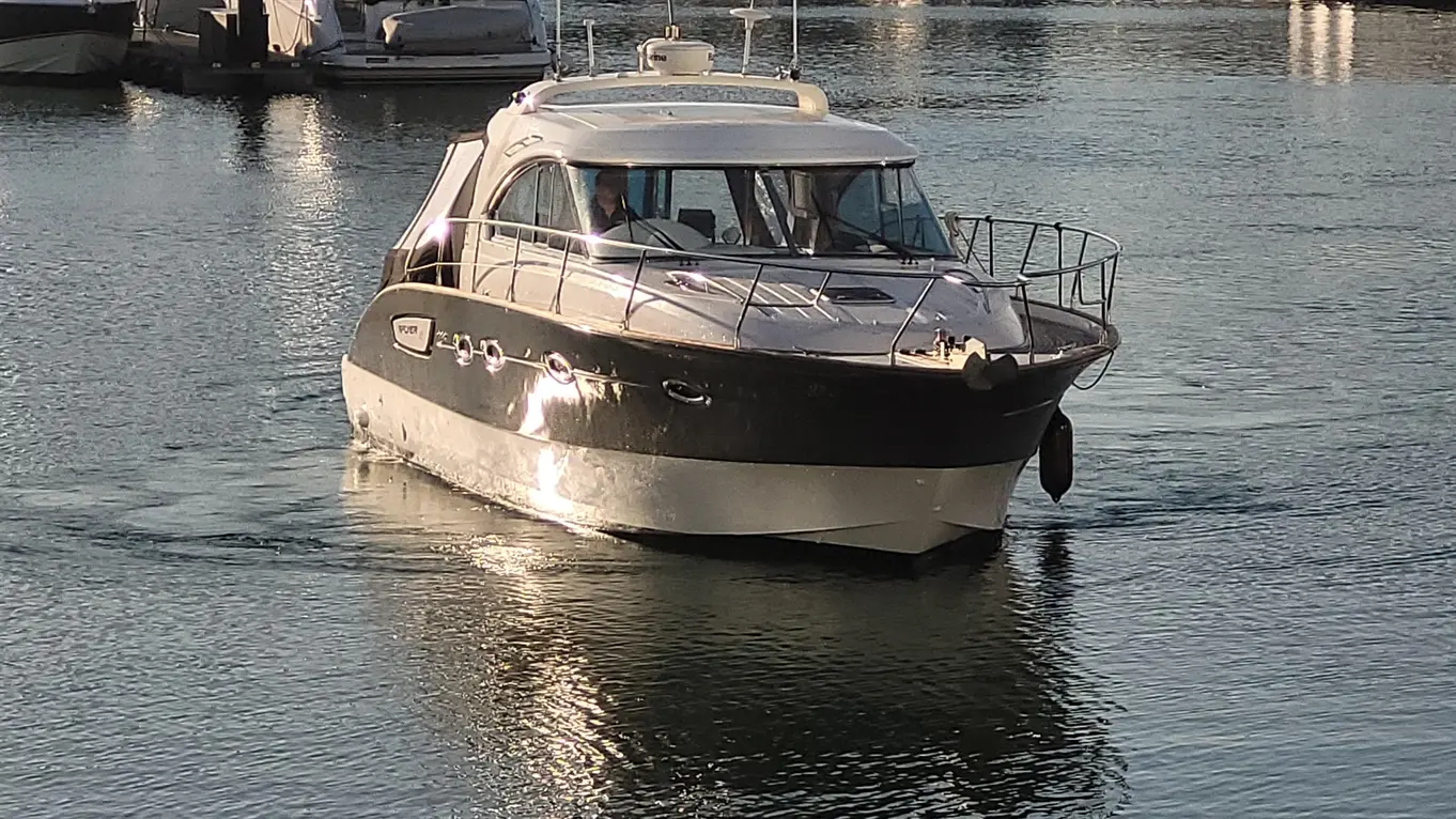 A sleek motorboat navigates through calm water in a marina, surrounded by other boats and reflecting sunlight.